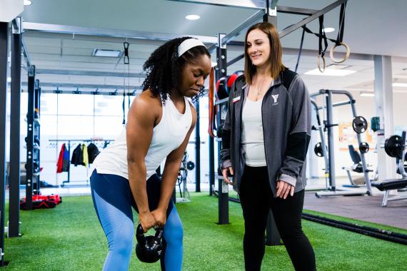 A woman in a white shirt and blue leggings lifts a black kettlebell with the guidance of a YMCA Trainer, on the turf fitness zone.