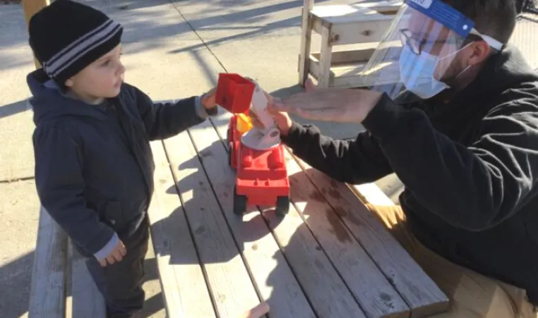 A young boy and Early Childhood Educator in PPE play at an outdoor picnic table with a red truck.