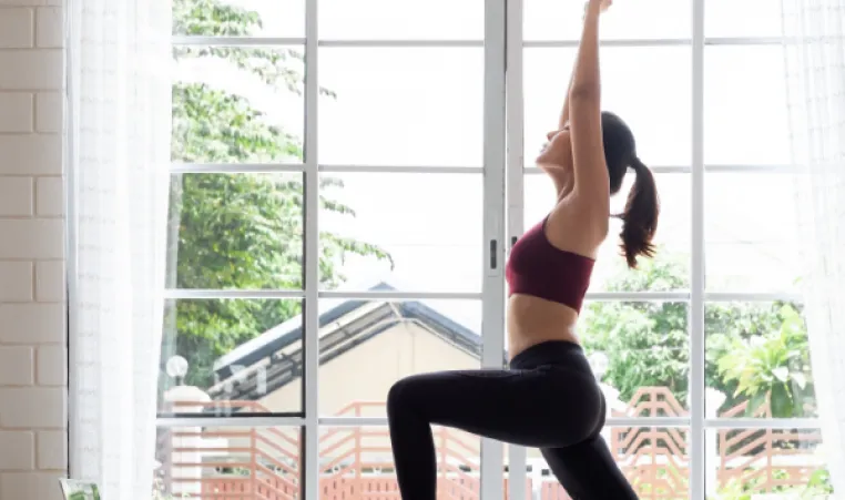 A woman practices yoga by a window at home, following an online workout on her laptop.