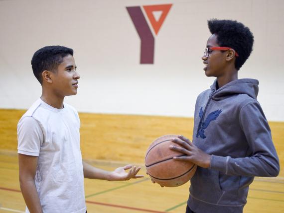 Two teenage boys talk while one holds a basketball in a YMCA gymnasium.