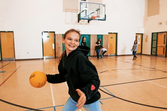 A teenage girl in jeans and a black sweatshirt laughs and gets ready to throw a ball during a game in a YMCA gymnasium.