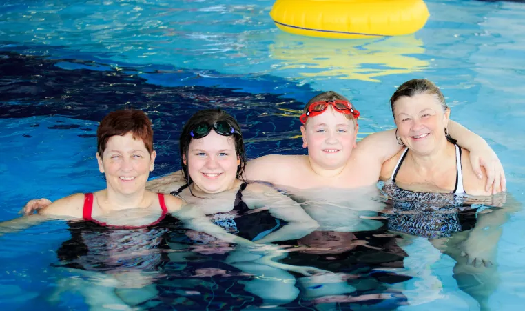 A family of four (two women, a girl and boy) smile and hug in a YMCA pool.