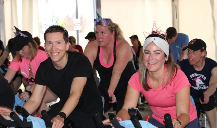 A group of people in pink and black shirts cycle on stationery bikes at a SWEAT for Strong Kids event.