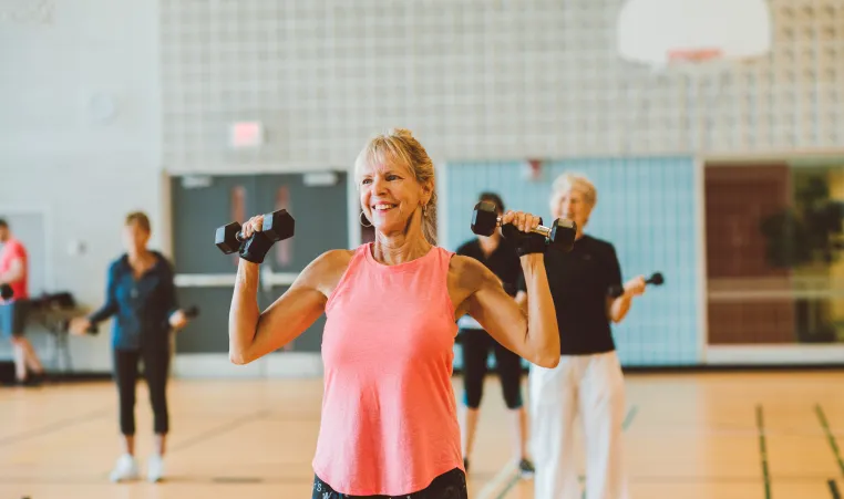 An older woman in a pink tank top smiles and lifts weights as part of a group fitness class.