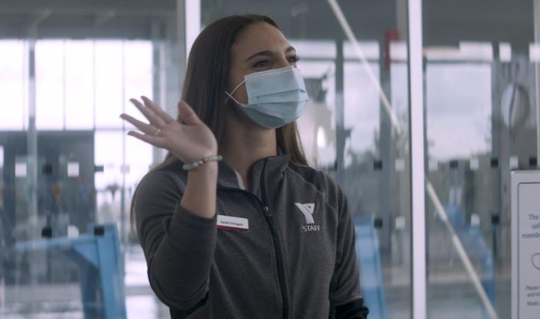 A YMCA staff member in a mask and Y shirt waves and greets members.