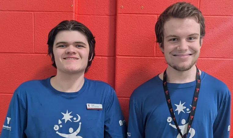 Two young men with light skin and short brown hair stand side by side in front of a red brick wall, smiling at the camera. They are both wearing matching blue t-shirts with a white logo featuring a stylized person, stars, and swirls. The man on the left has a name tag that reads 'Noah' and 'Project Search Team.' The man on the right wears a black lanyard with red 'Y' logos.