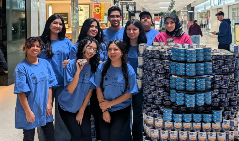 A group of nine young volunteers wearing matching blue t-shirts with "The Inn of The Good Shepherd" logo stand together in a shopping mall, smiling at the camera. They are positioned next to a large, neatly stacked display of canned food, primarily tuna and chickpeas. One volunteer in the front row holds up a can. In the background, shoppers walk through the mall, and a sign for the YMCA Canned Food Drive is visible.