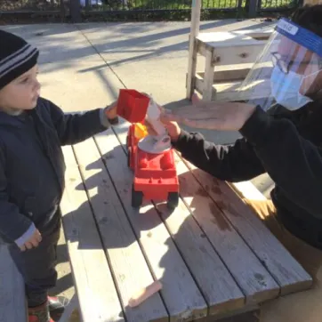 A young boy and Early Childhood Educator in PPE play at an outdoor picnic table with a red truck.