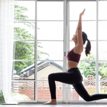 A woman practices yoga by a window at home, following an online workout on her laptop.