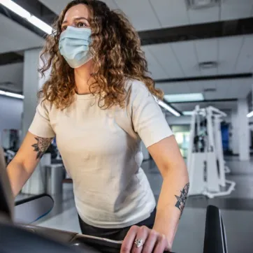 A woman with curly hair and tattoos on her arm, wearing a mask and white t-shirt, works out on a treadmill at the Y.