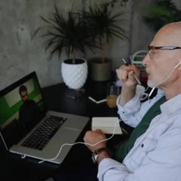 A man in a white button-up shirt with glasses and headphones sits at a computer desk while having a video call with another man on his laptop.