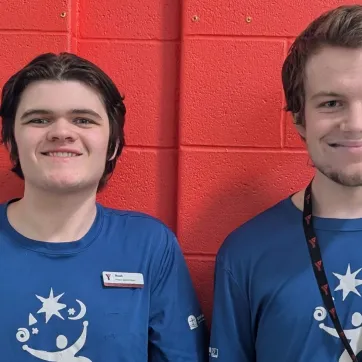 Two young men with light skin and short brown hair stand side by side in front of a red brick wall, smiling at the camera. They are both wearing matching blue t-shirts with a white logo featuring a stylized person, stars, and swirls. The man on the left has a name tag that reads 'Noah' and 'Project Search Team.' The man on the right wears a black lanyard with red 'Y' logos.