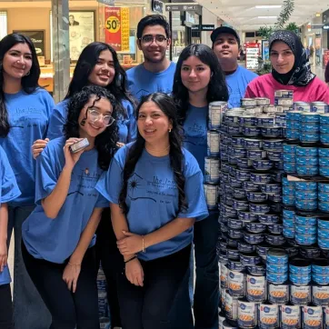 A group of nine young volunteers wearing matching blue t-shirts with "The Inn of The Good Shepherd" logo stand together in a shopping mall, smiling at the camera. They are positioned next to a large, neatly stacked display of canned food, primarily tuna and chickpeas. One volunteer in the front row holds up a can. In the background, shoppers walk through the mall, and a sign for the YMCA Canned Food Drive is visible.